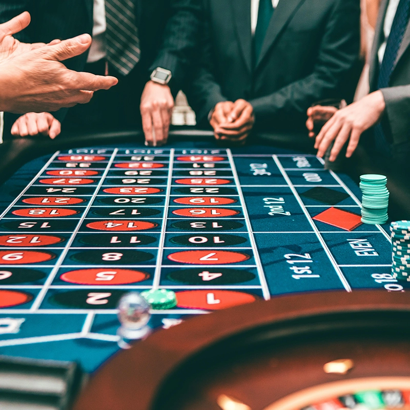 People gathered around a roulette table in a casino, with red and black numbered spaces and poker chips visible.